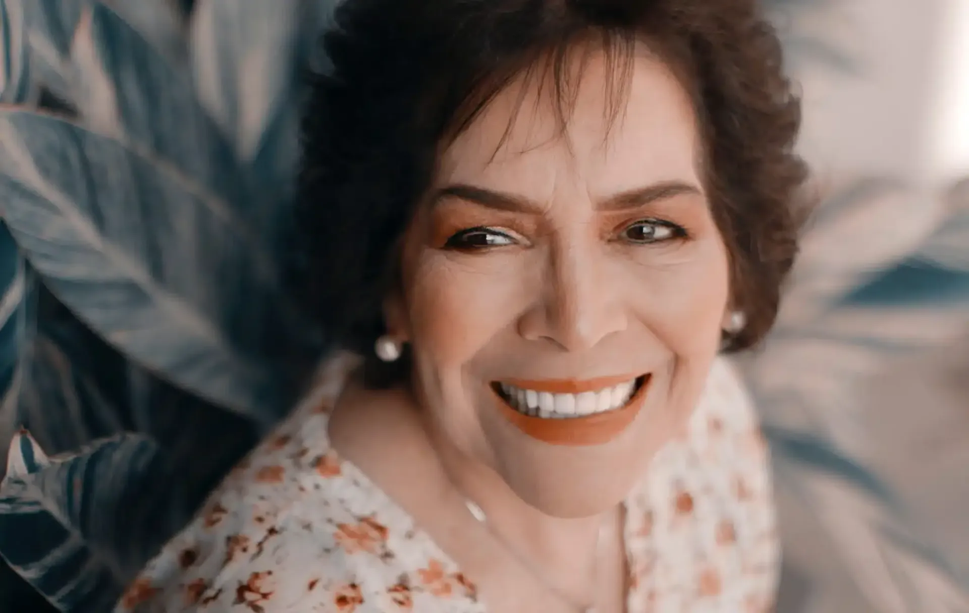 Smiling older woman with dark hair, wearing a floral top, surrounded by leafy plants.