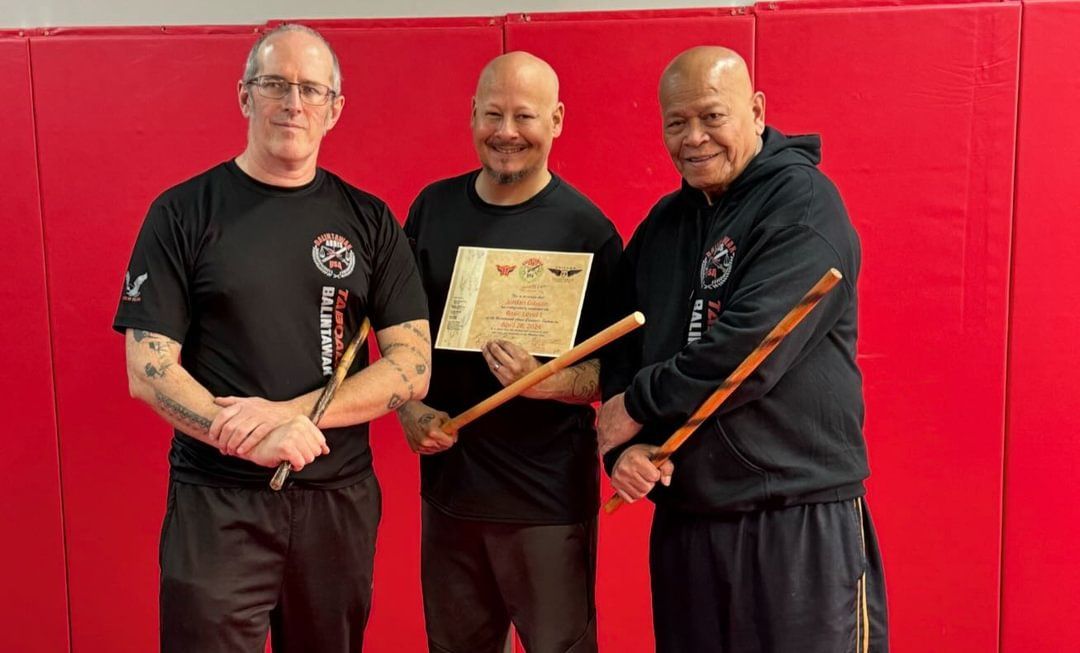 Three men are standing next to each other in a gym holding wooden sticks.