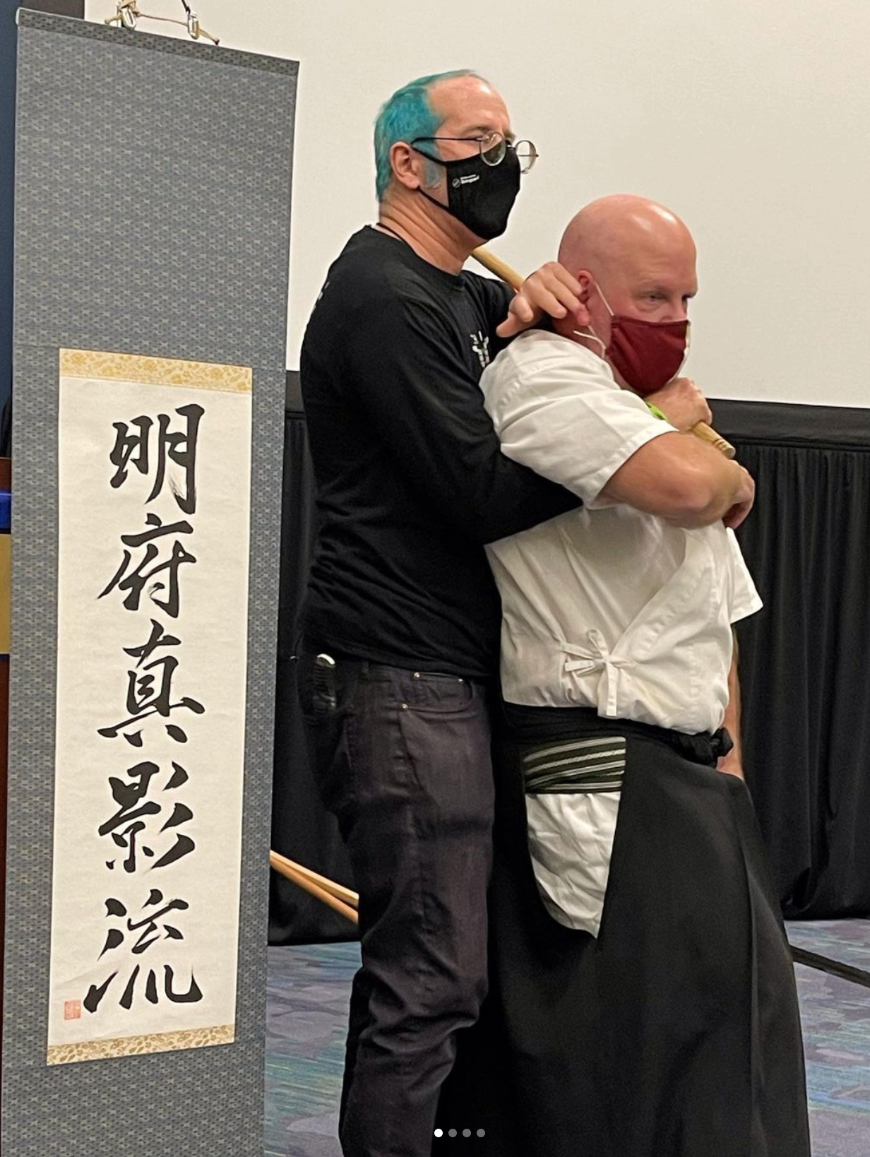 Two men wearing masks are hugging each other in front of a wall with chinese writing on it.