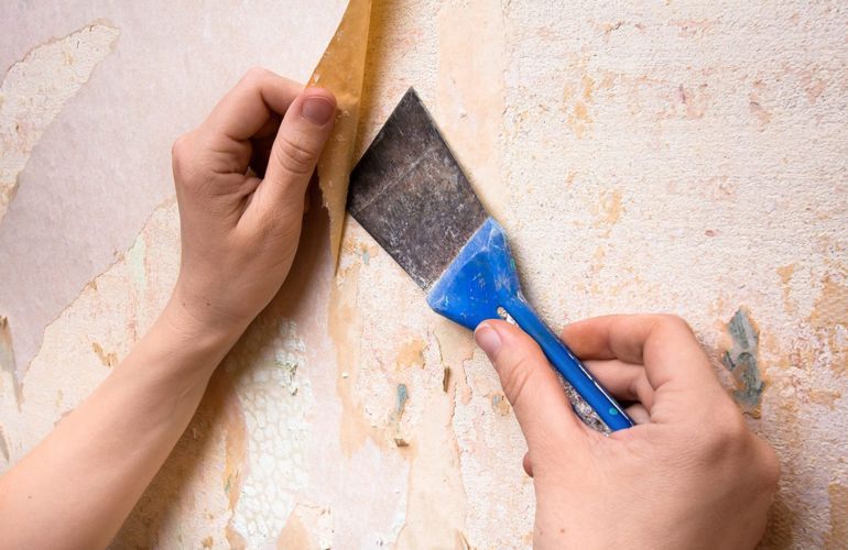 A person is removing wallpaper from a wall with a spatula.