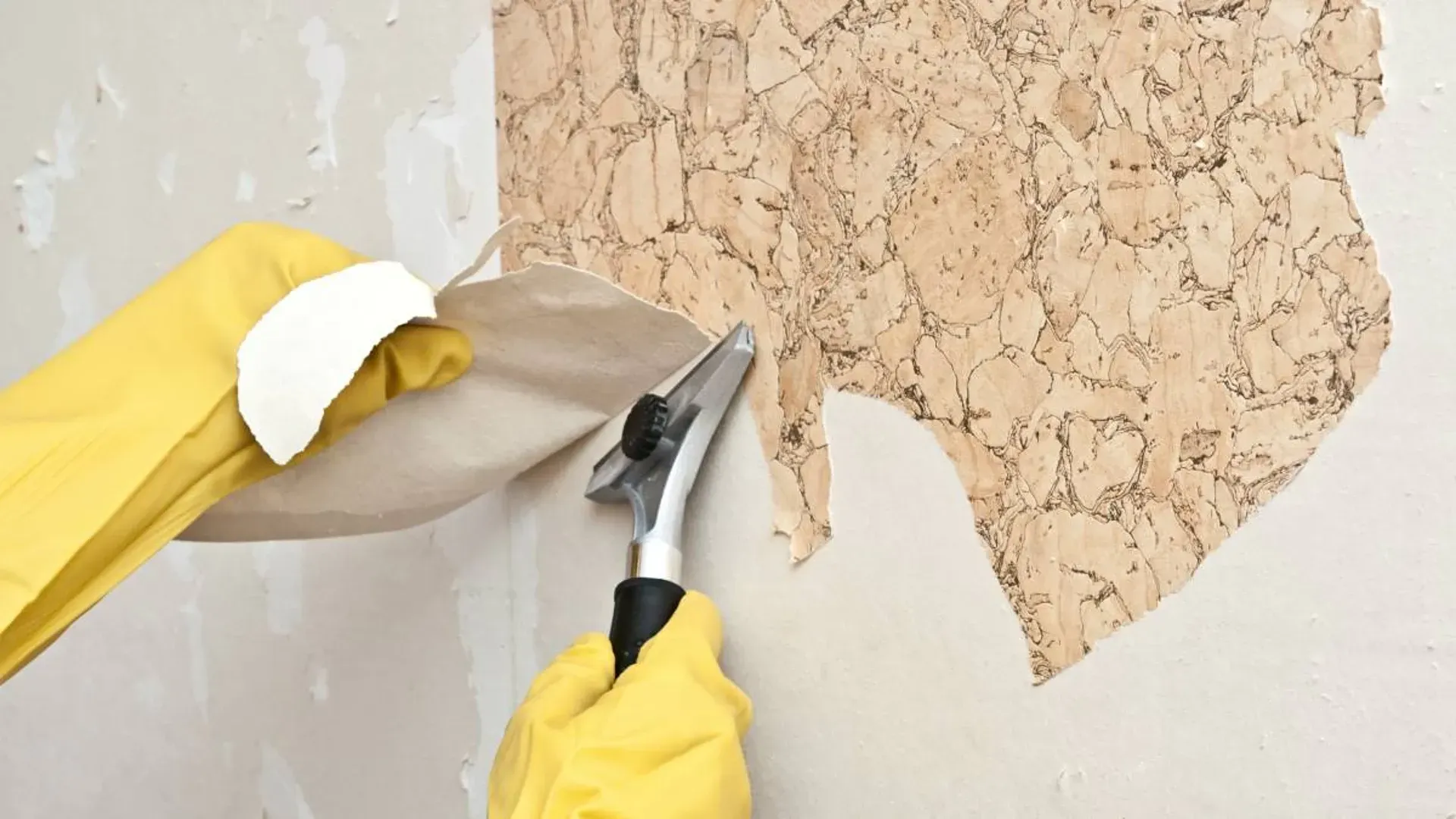 A person wearing yellow gloves is removing wallpaper from a wall with a spatula.