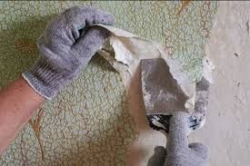 A person wearing gloves is removing wallpaper from a wall with a spatula.