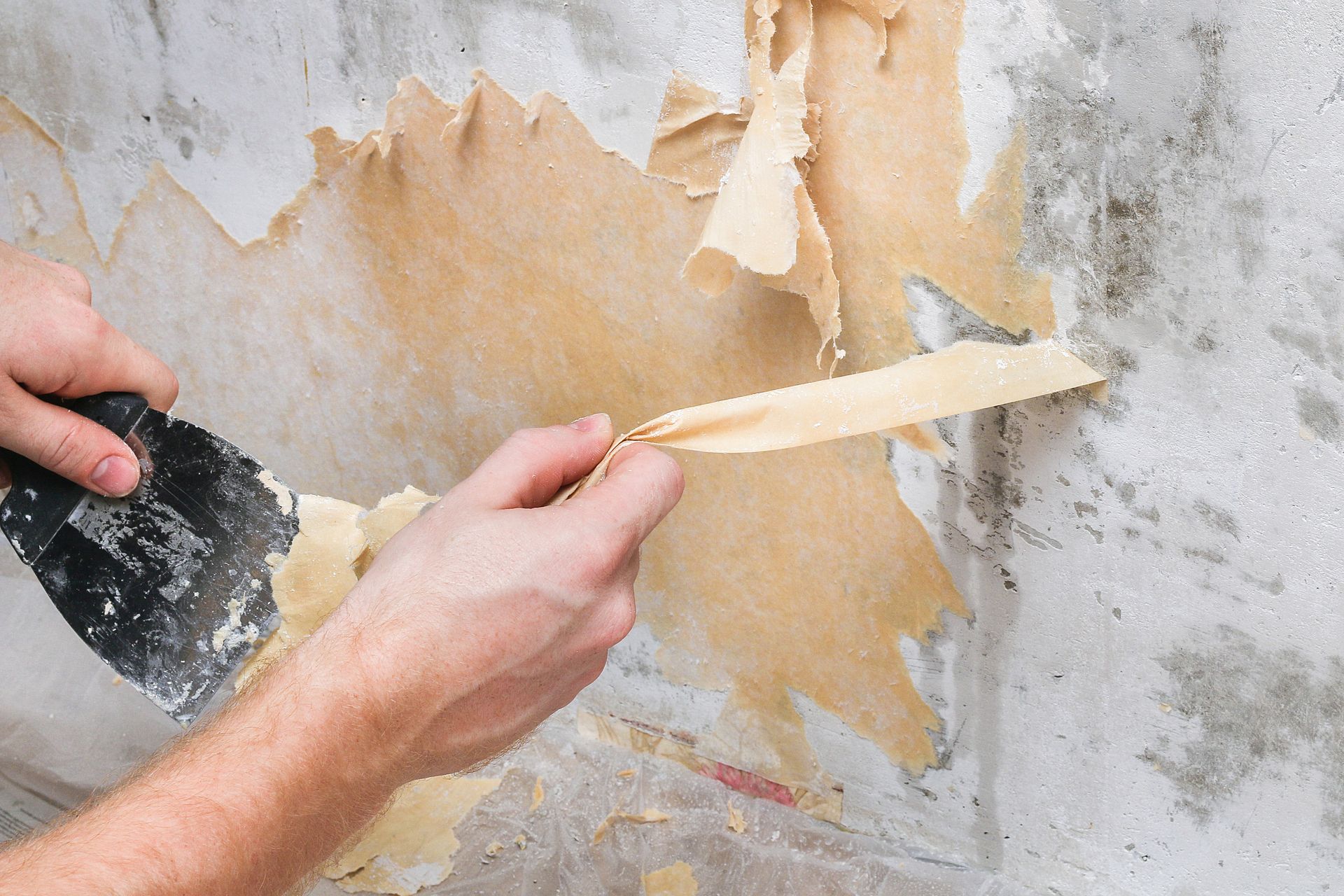 A person is peeling wallpaper from a wall with a spatula.