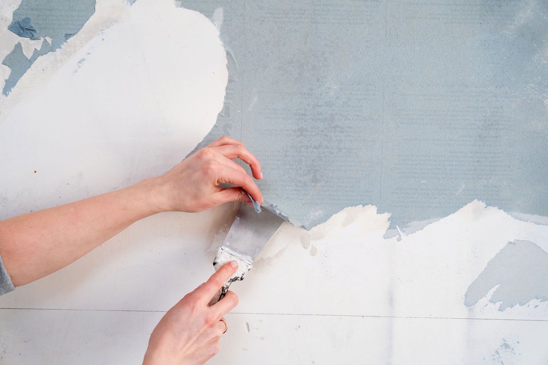 A person is peeling wallpaper from a wall with a spatula.
