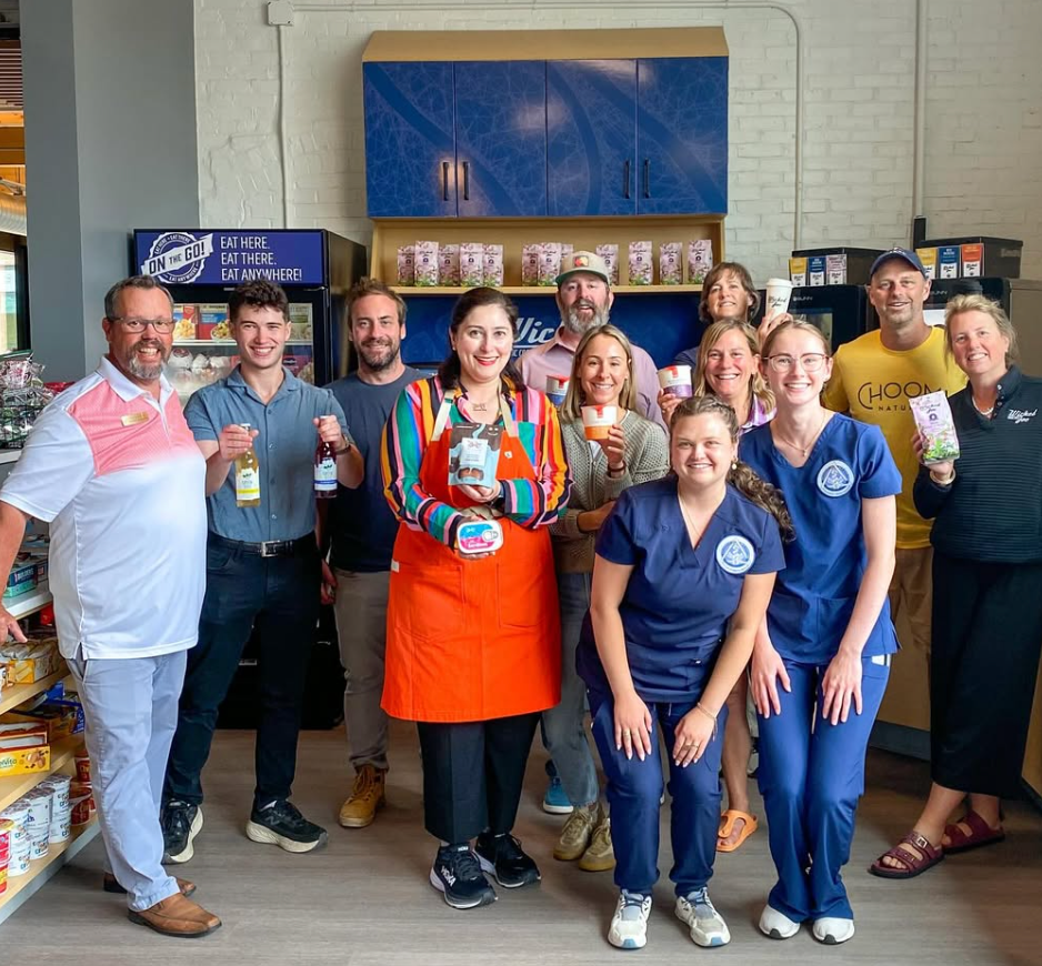 Group of people posing inside a shop with food products on shelves. Some hold food or drink, smiling.