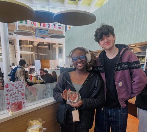 Two people smiling, holding plates with burgers and drinks; a room with others seated at tables.