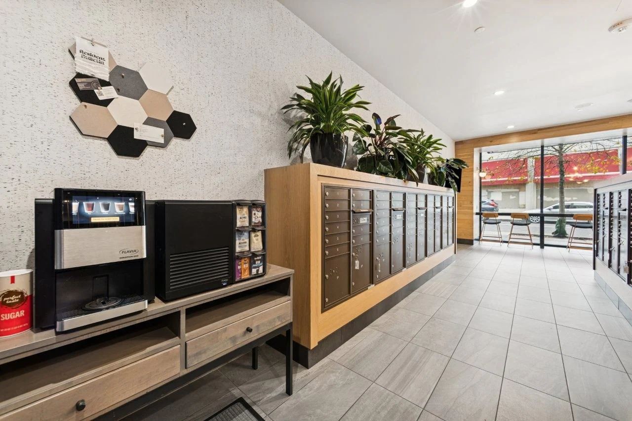Lobby with mailboxes, coffee machine, and plants against a textured wall and large windows.