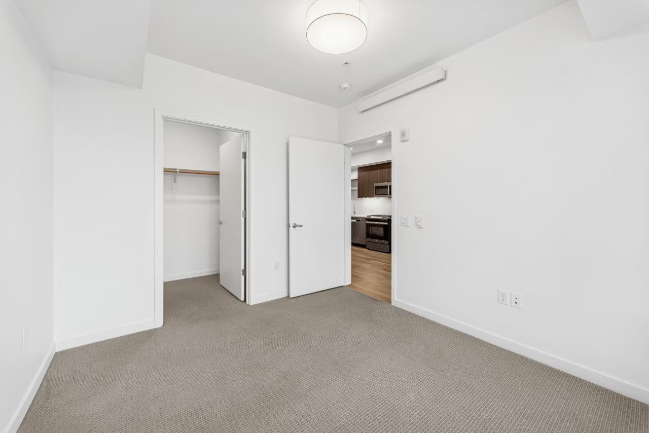 Empty bedroom with a closet and a doorway leading to a kitchen. Beige carpet and white walls.