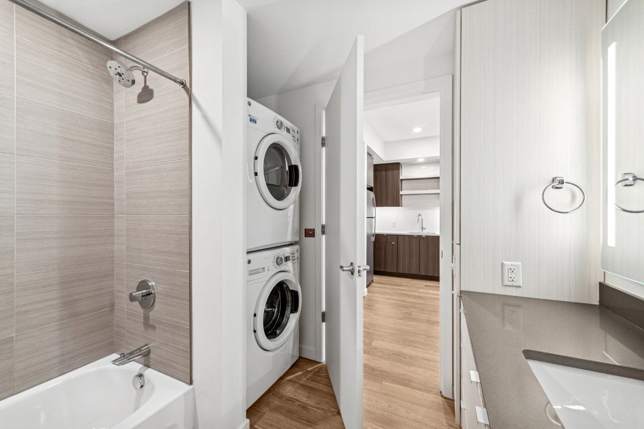 Bathroom with stacked washer/dryer, shower, vanity, and open doorway leading to a kitchen.