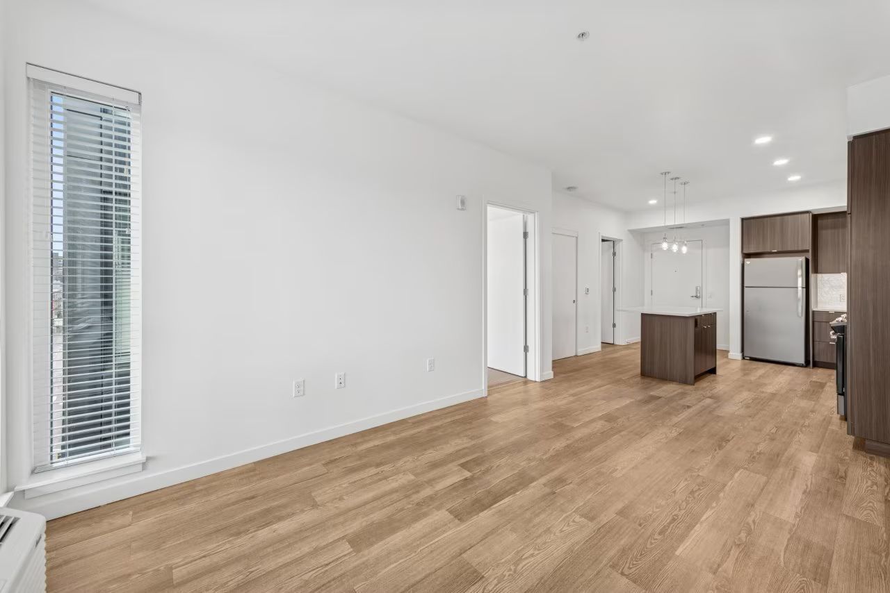 Empty modern apartment with hardwood floors, kitchen, and large window with blinds.