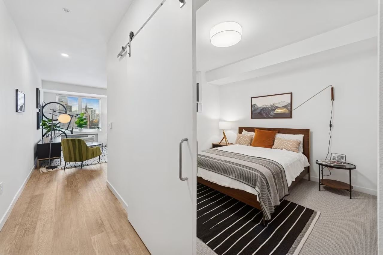 Bedroom interior with desk area visible through open sliding door; light wood floors, white walls, and neutral decor.