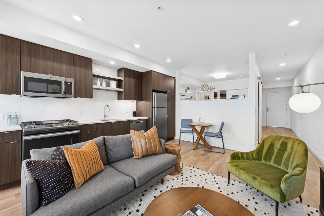 Modern apartment living room with kitchen, sofa, green chair, round table, rug.