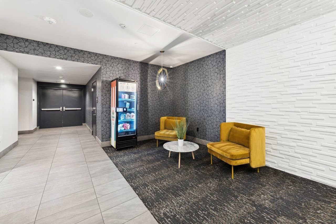 Lobby with two yellow chairs, a vending machine, and a small table. Black and white patterned carpet.