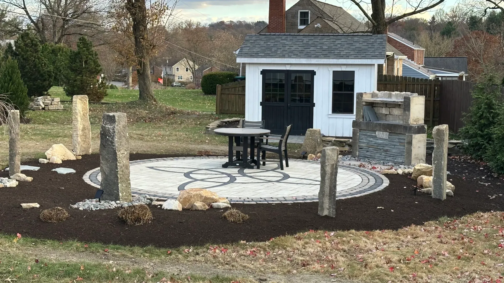 Memorial garden with stone markers, circular brick patio, bench, and small white shed in the background