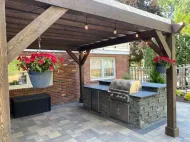 Backyard patio with wooden pergola, stone outdoor kitchen, grill, and hanging red flowers