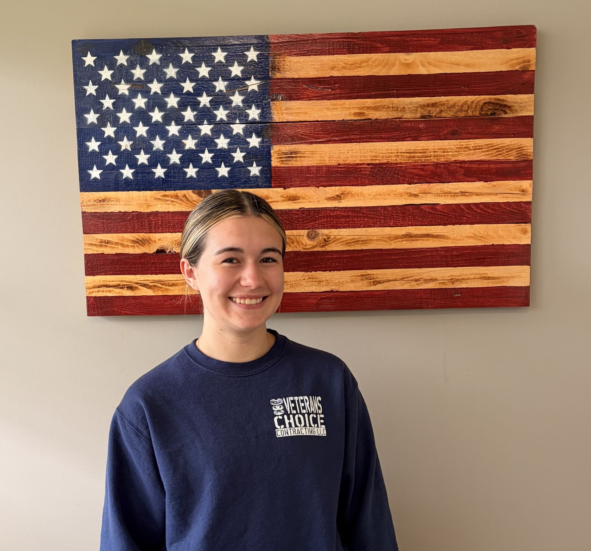 Person smiling in a navy sweatshirt before a wooden American flag wall hanging