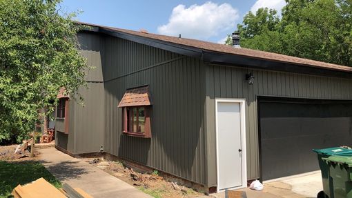Exterior of a house and garage with olive green siding, brown roof and trim, and white door.