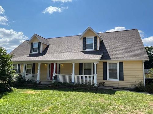 Yellow house with a wraparound porch, two dormers, and black shutters, under a blue sky.