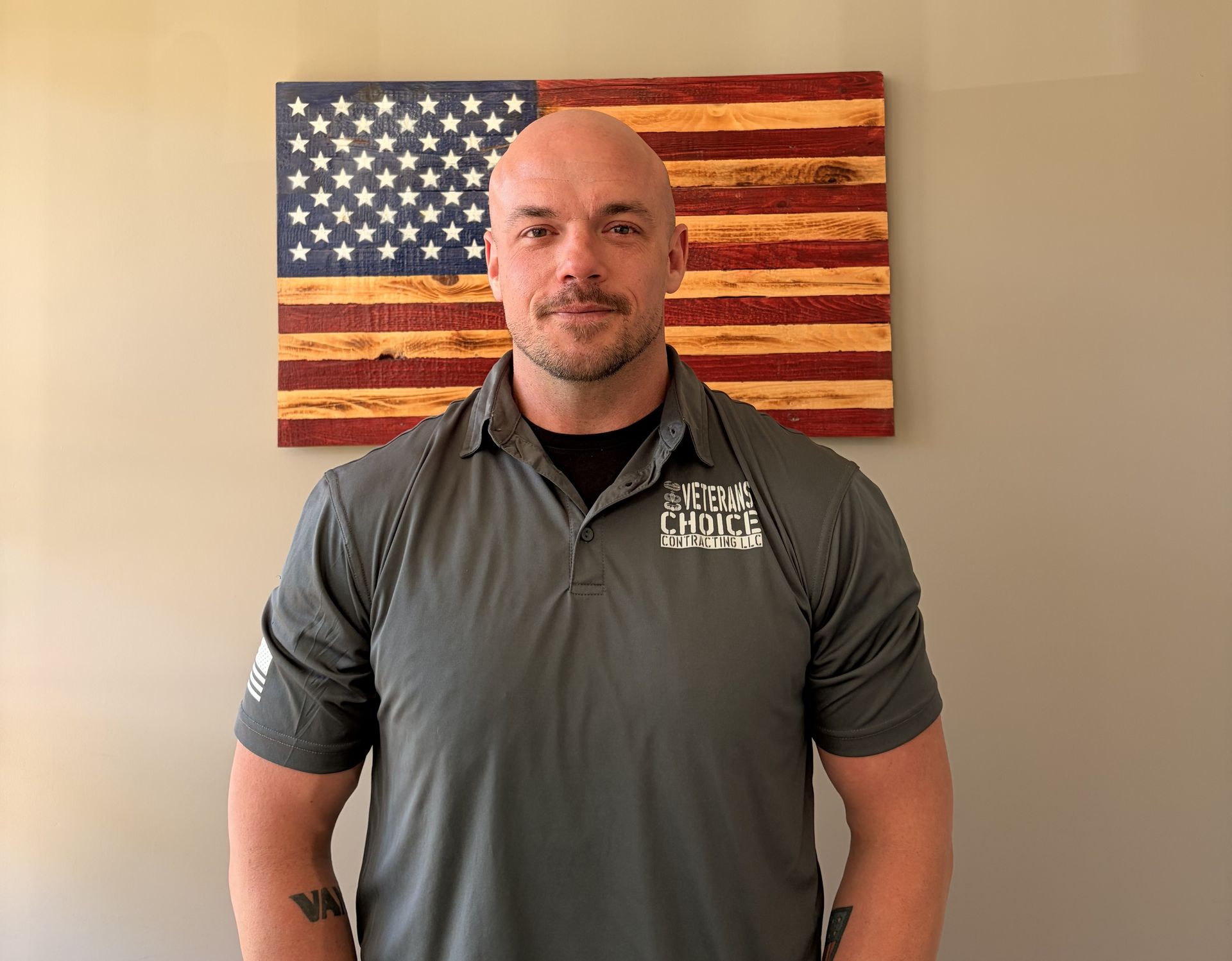 Man standing indoors before a wooden American flag sign, wearing a gray polo shirt