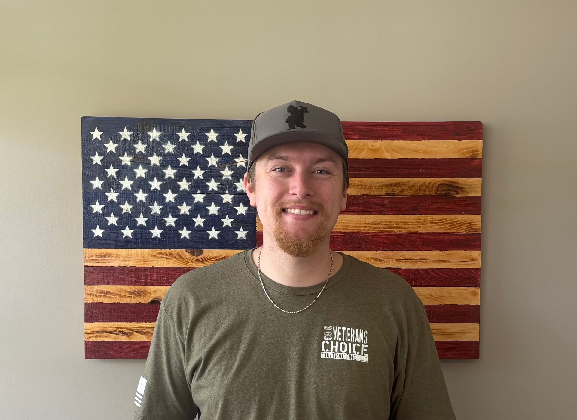 Man with folded arms standing before a painted American flag backdrop, wearing an olive T-shirt.