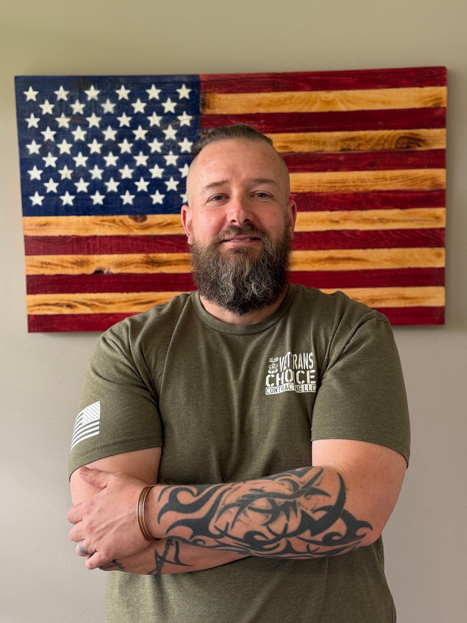 Man with folded arms standing before a painted American flag backdrop, wearing an olive T-shirt.