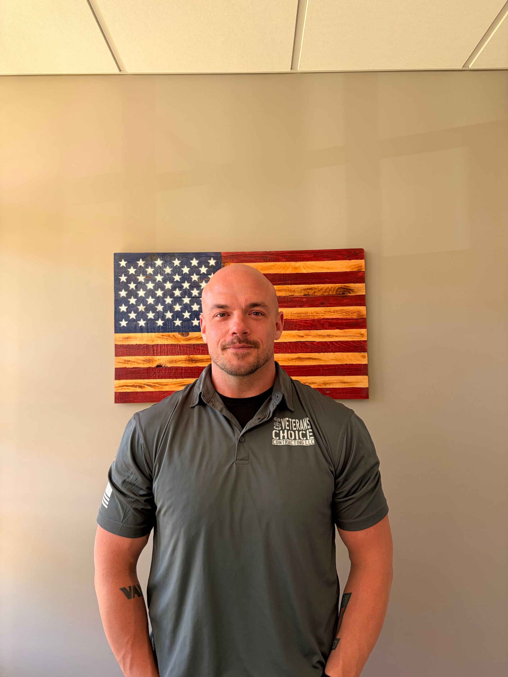 Man with folded arms standing before a painted American flag backdrop, wearing an olive T-shirt.