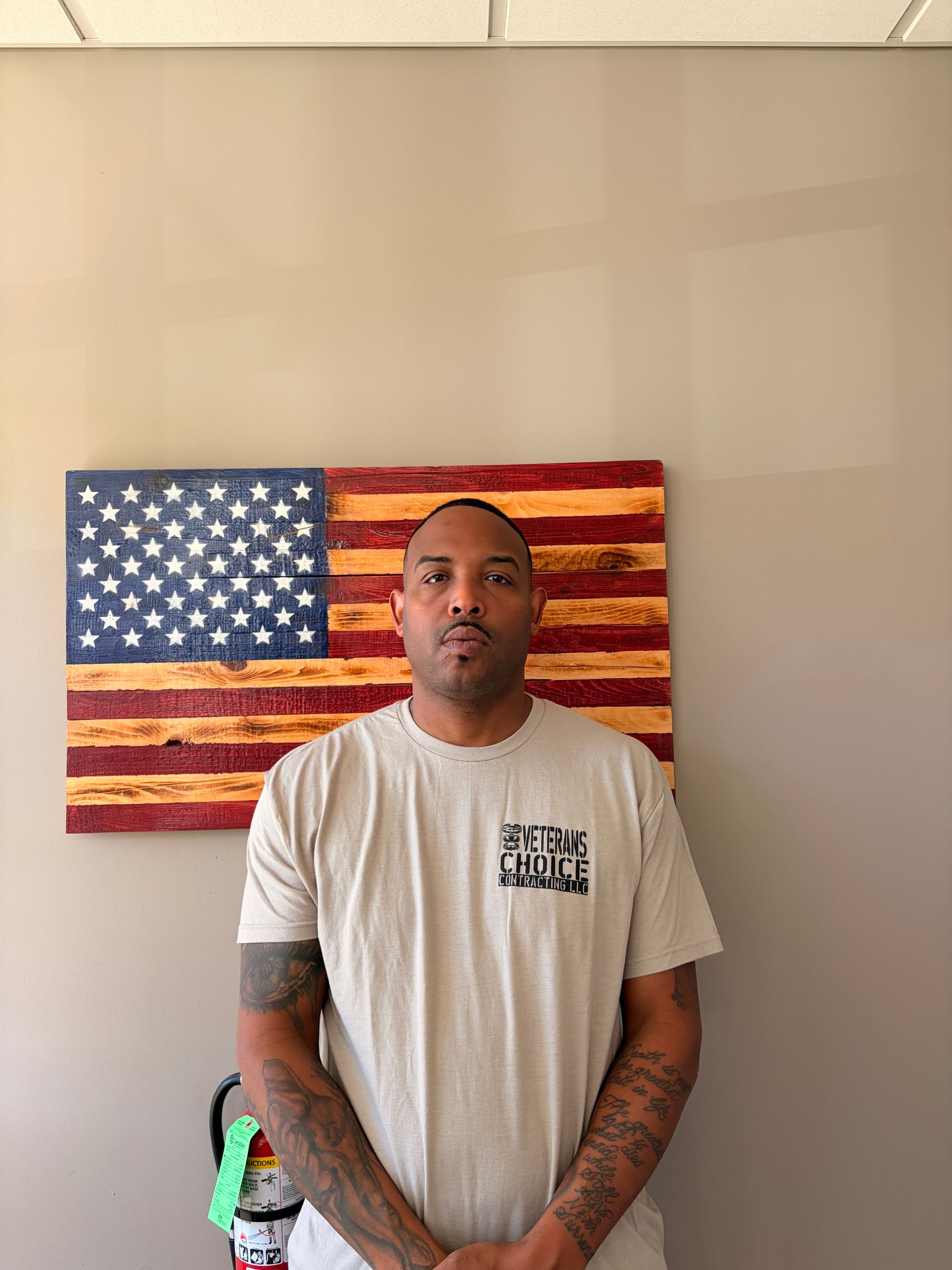 Man with folded arms standing before a painted American flag backdrop, wearing an olive T-shirt.