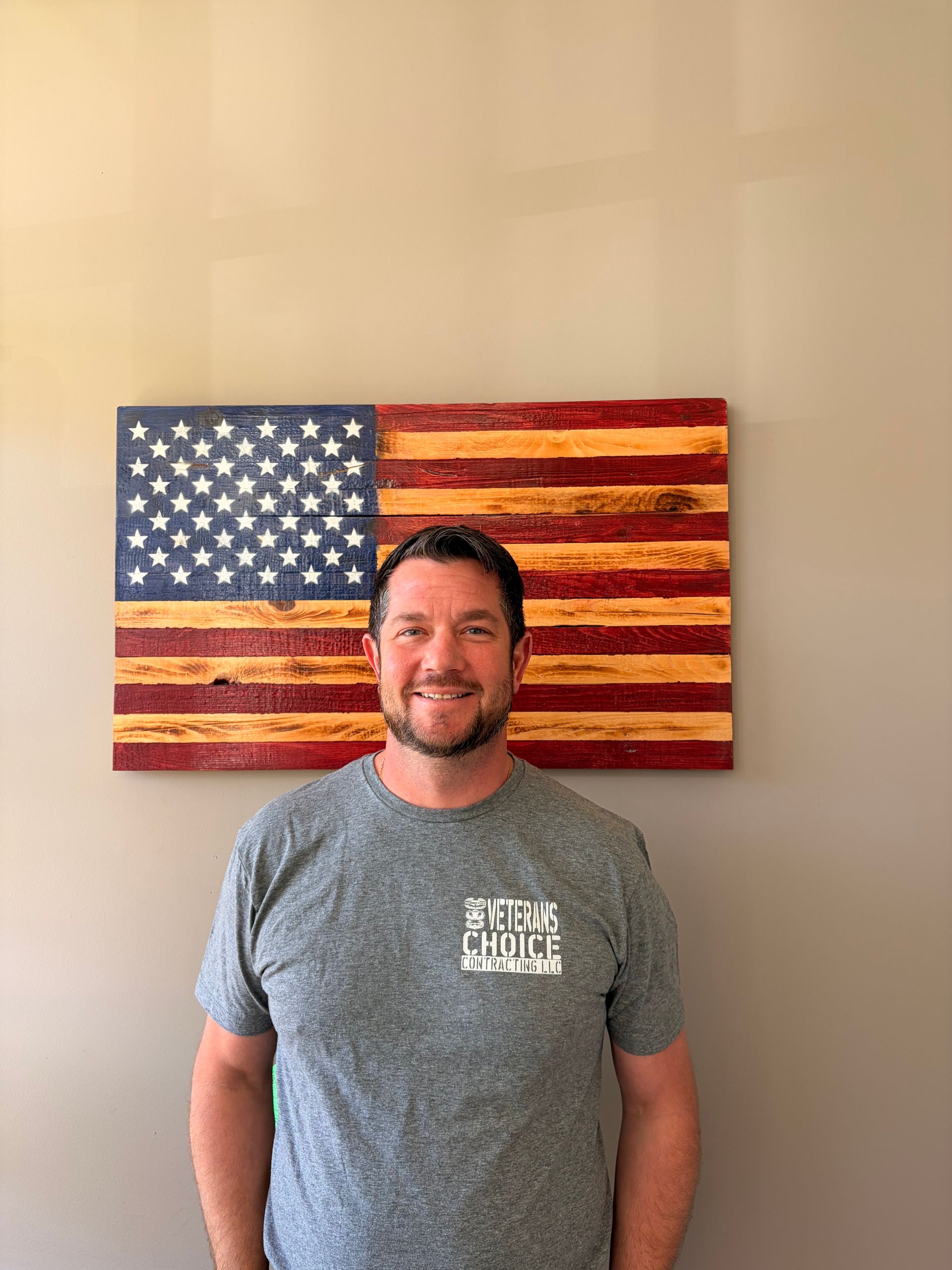 Man with folded arms standing before a painted American flag backdrop, wearing an olive T-shirt.