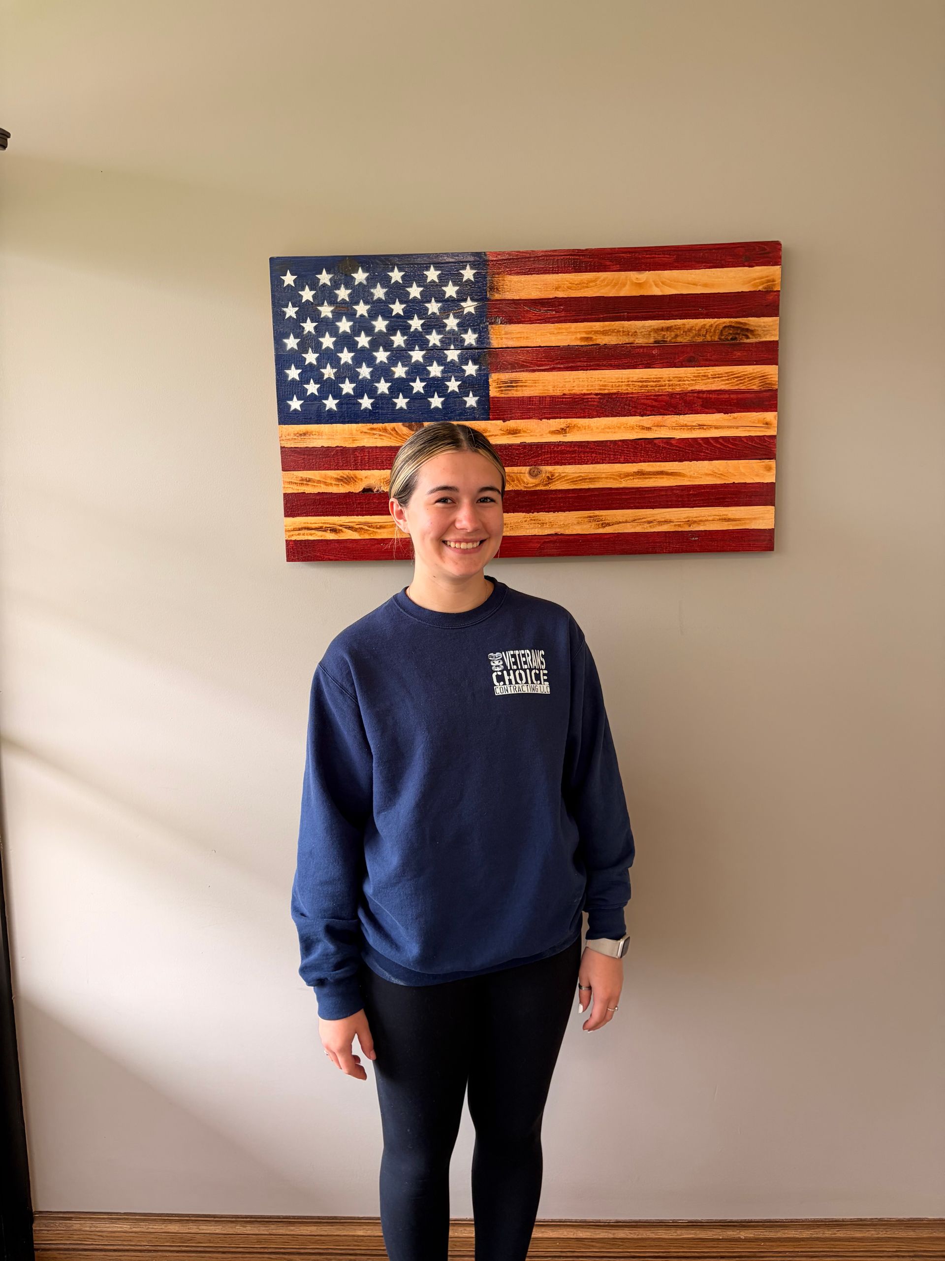 Smiling person standing before a rustic wooden American flag on a beige wall