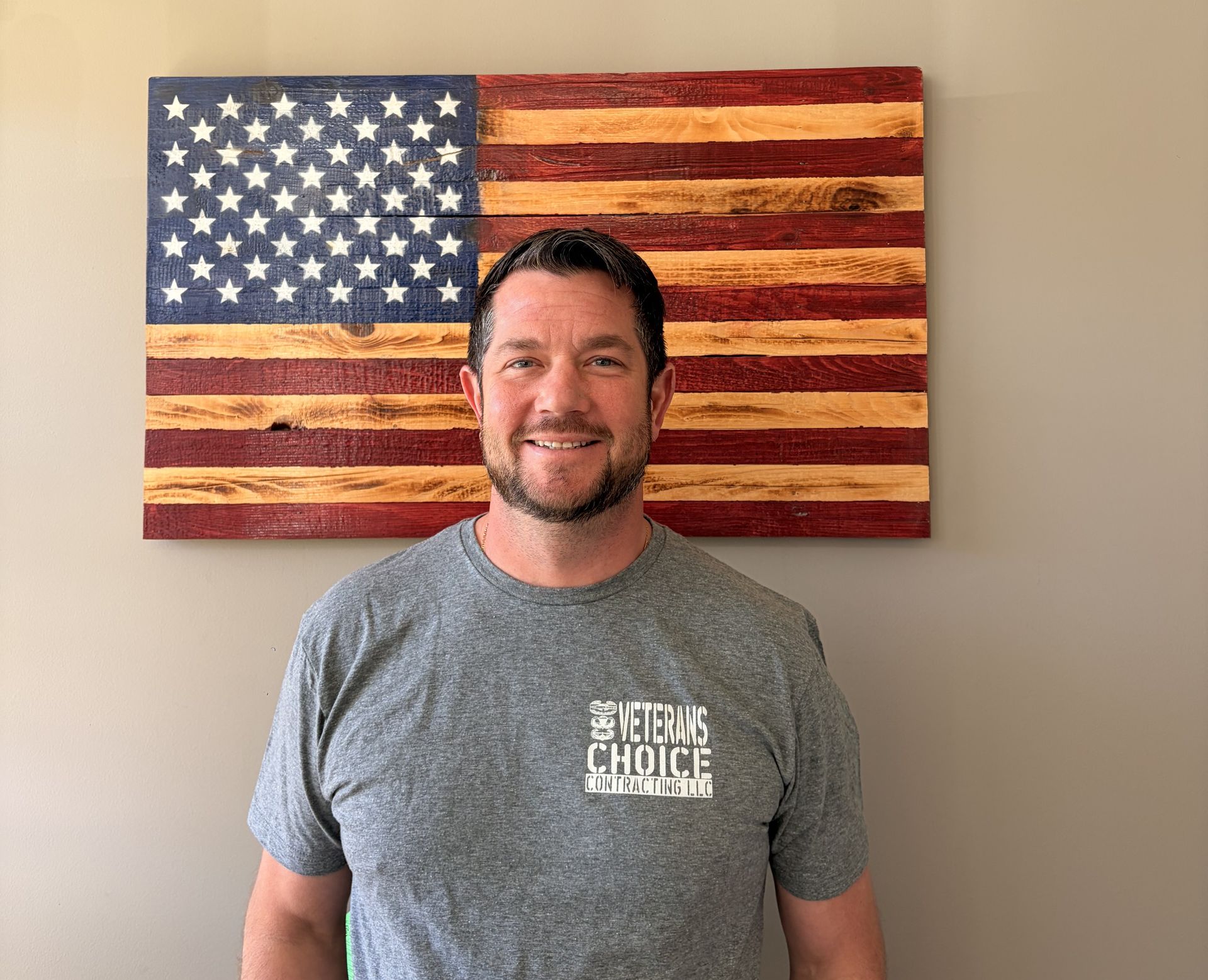 Man in black polo shirt stands in front of a wooden American flag.