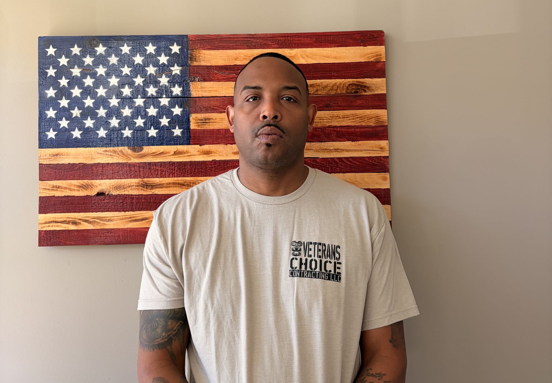 Man standing indoors before a distressed American flag wall art, wearing a beige T-shirt.