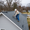 Worker installing shingles on a gray roof with a hammer, viewed from above in a suburban neighborhood