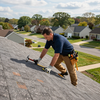 Worker installing shingles on a rooftop with tools, suburban houses visible in the background
