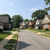 Quiet suburban street with brick and siding houses, sidewalks, and trees under a clear blue sky