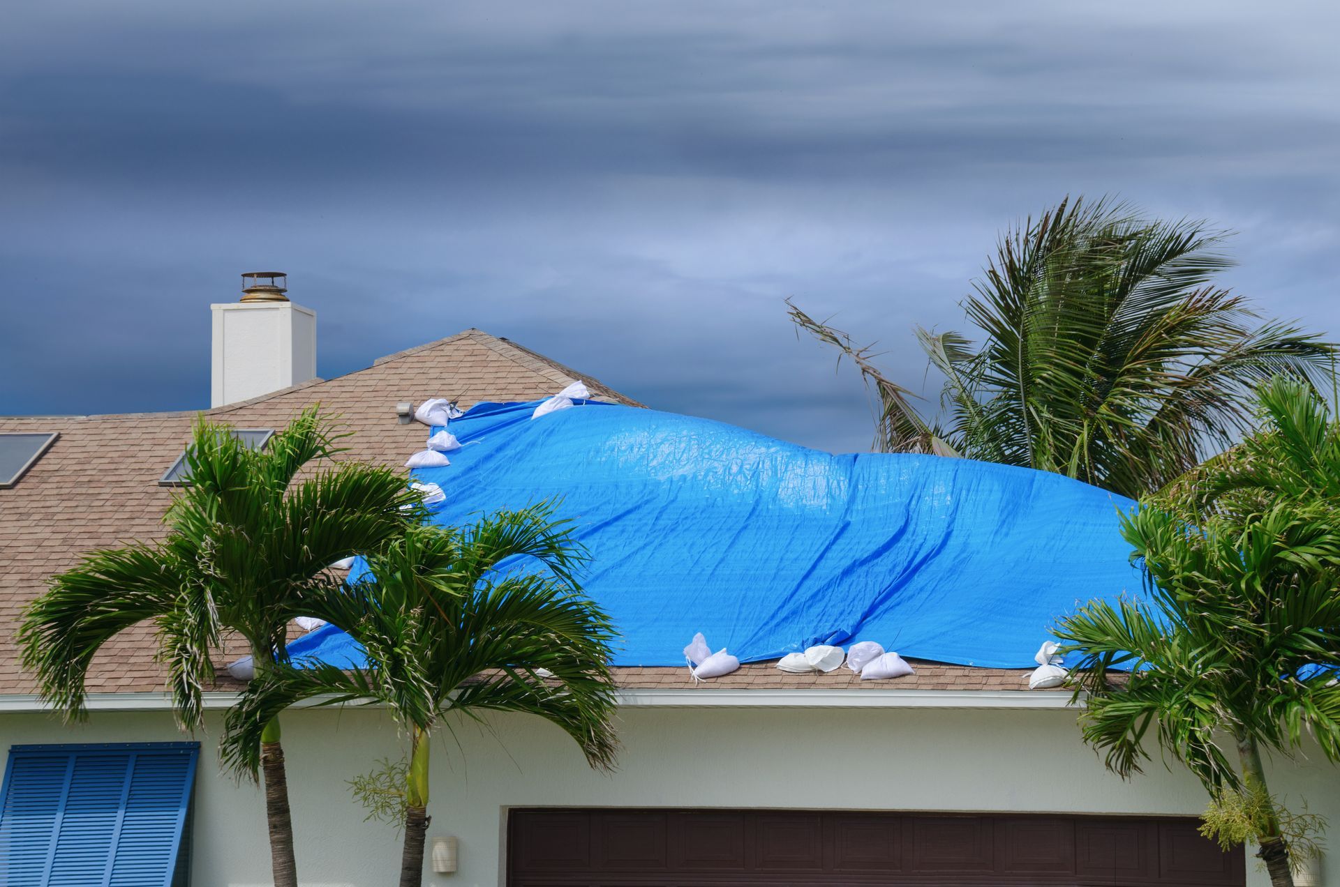House roof covered with a bright blue tarp after storm damage, with palm trees in front