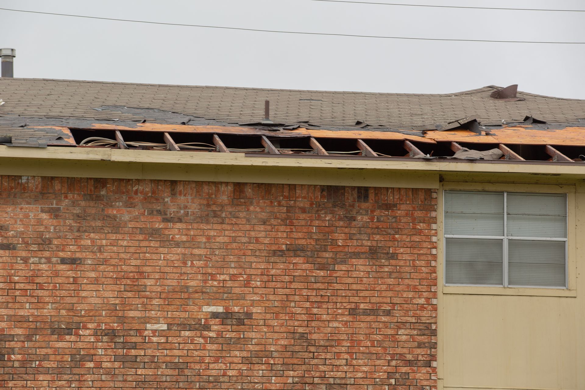 Damaged brick building roof with missing panels and exposed beams beneath a gray sky