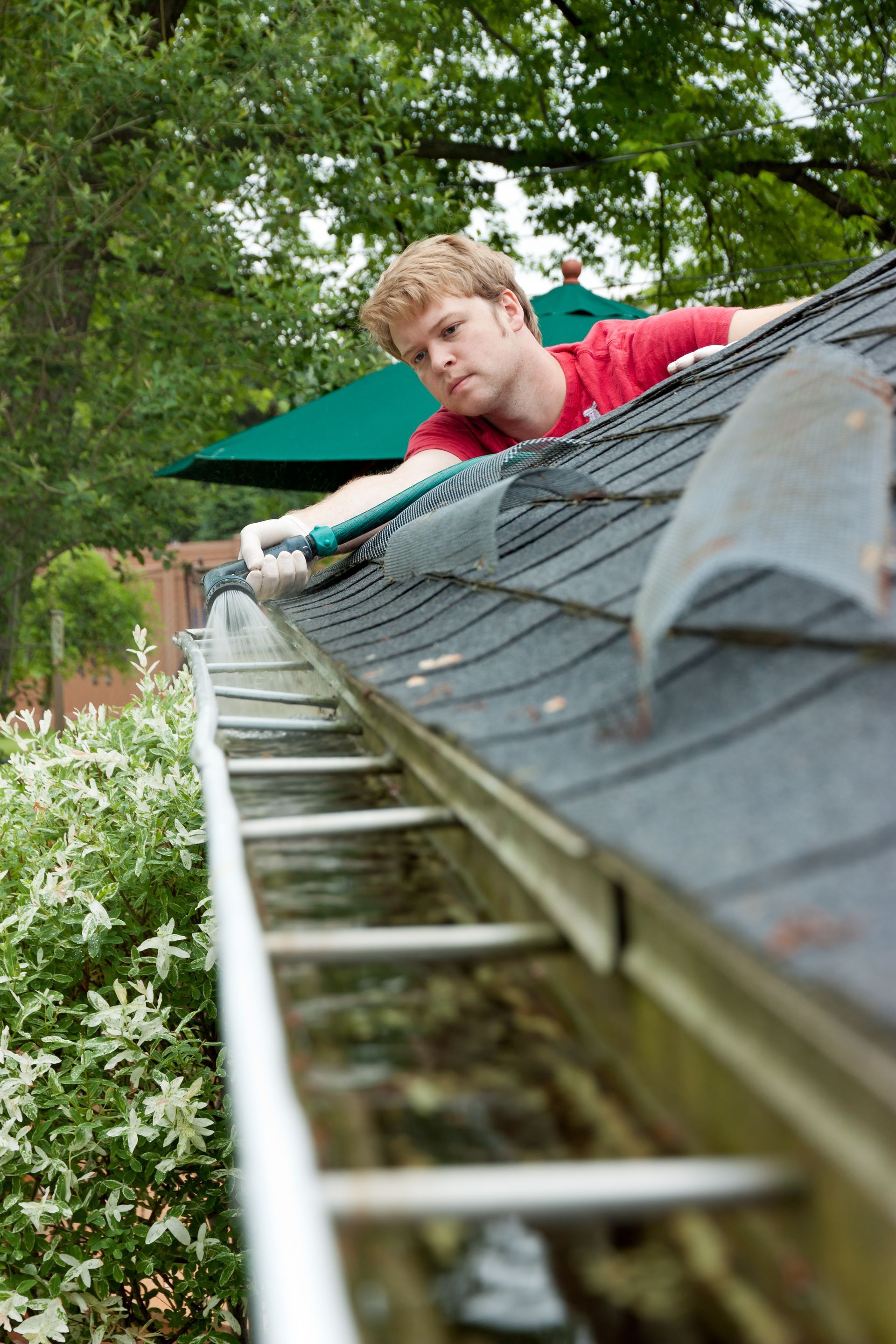 Person cleaning a roof gutter with a hose, standing on a ladder beside a house