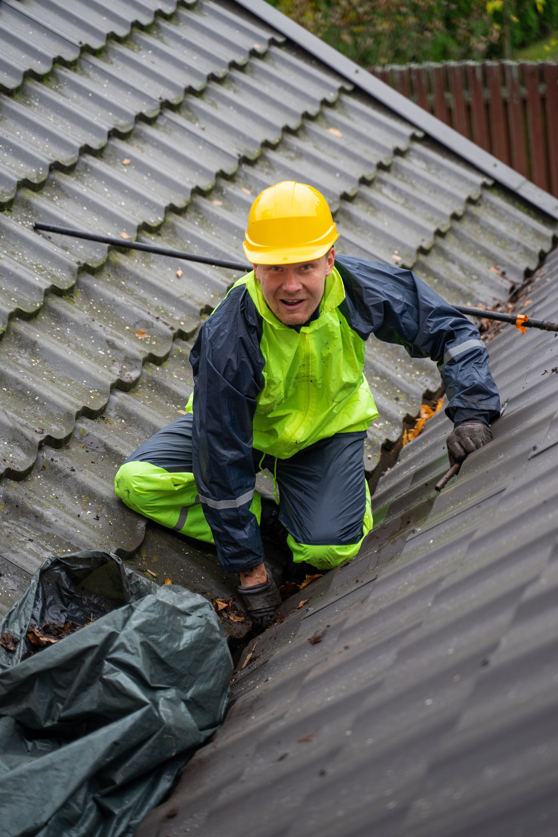 Worker in yellow hard hat repairing a wet roof in rain, kneeling beside damaged shingles.
