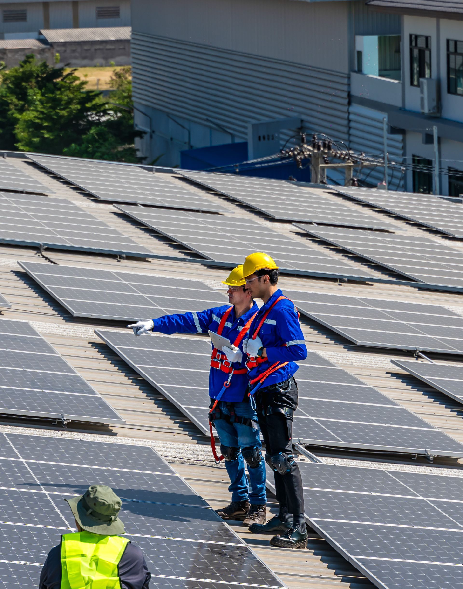 Workers inspecting solar panels on a rooftop, one pointing while wearing hard hats and safety gear