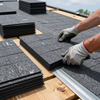 Gloved hands installing dark roof shingles on a wooden roof deck outdoors