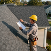 Roof inspector in yellow hard hat examining a shingled roof with a clipboard outdoors