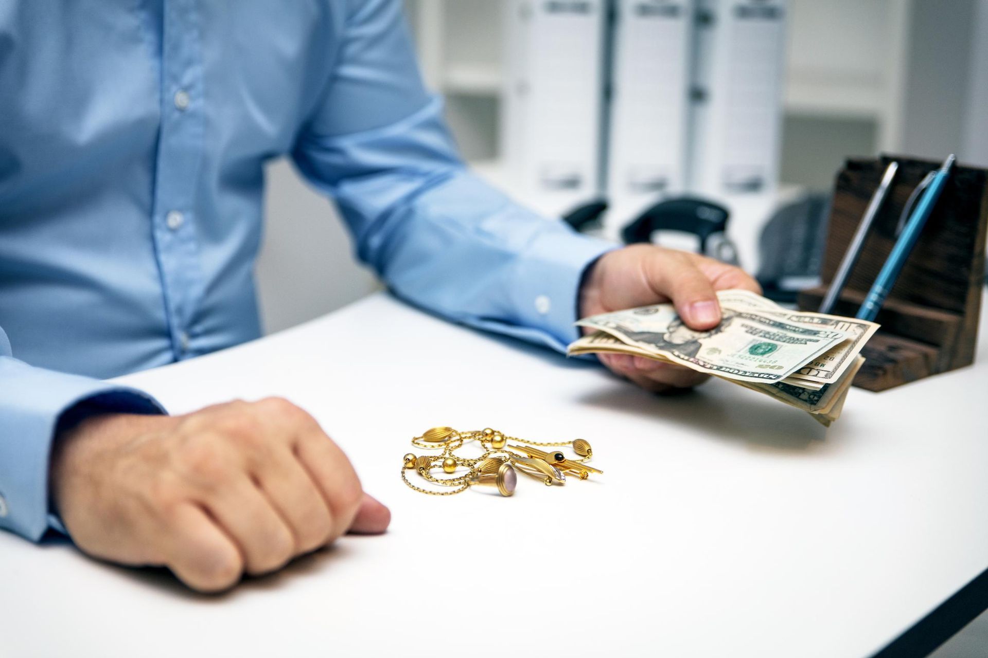 A man is sitting at a desk holding a stack of money and gold jewelry.
