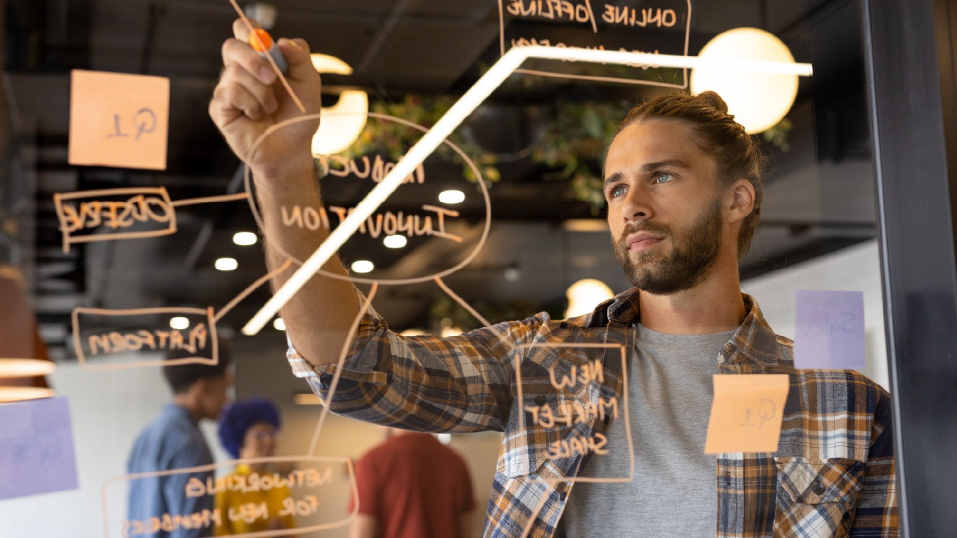 A person with a beard and tied hair writes on a glass board with notes and diagrams in a bright office workspace.