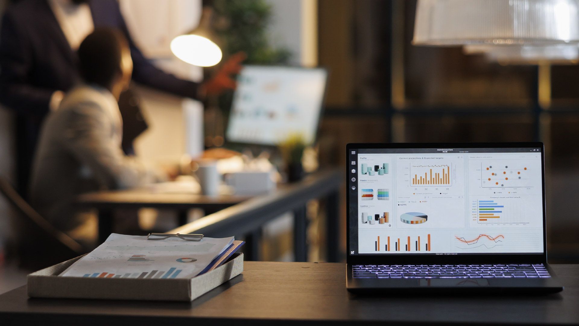 A laptop displaying data charts sits on a desk in the foreground, with blurred office workers collaborating in the back.