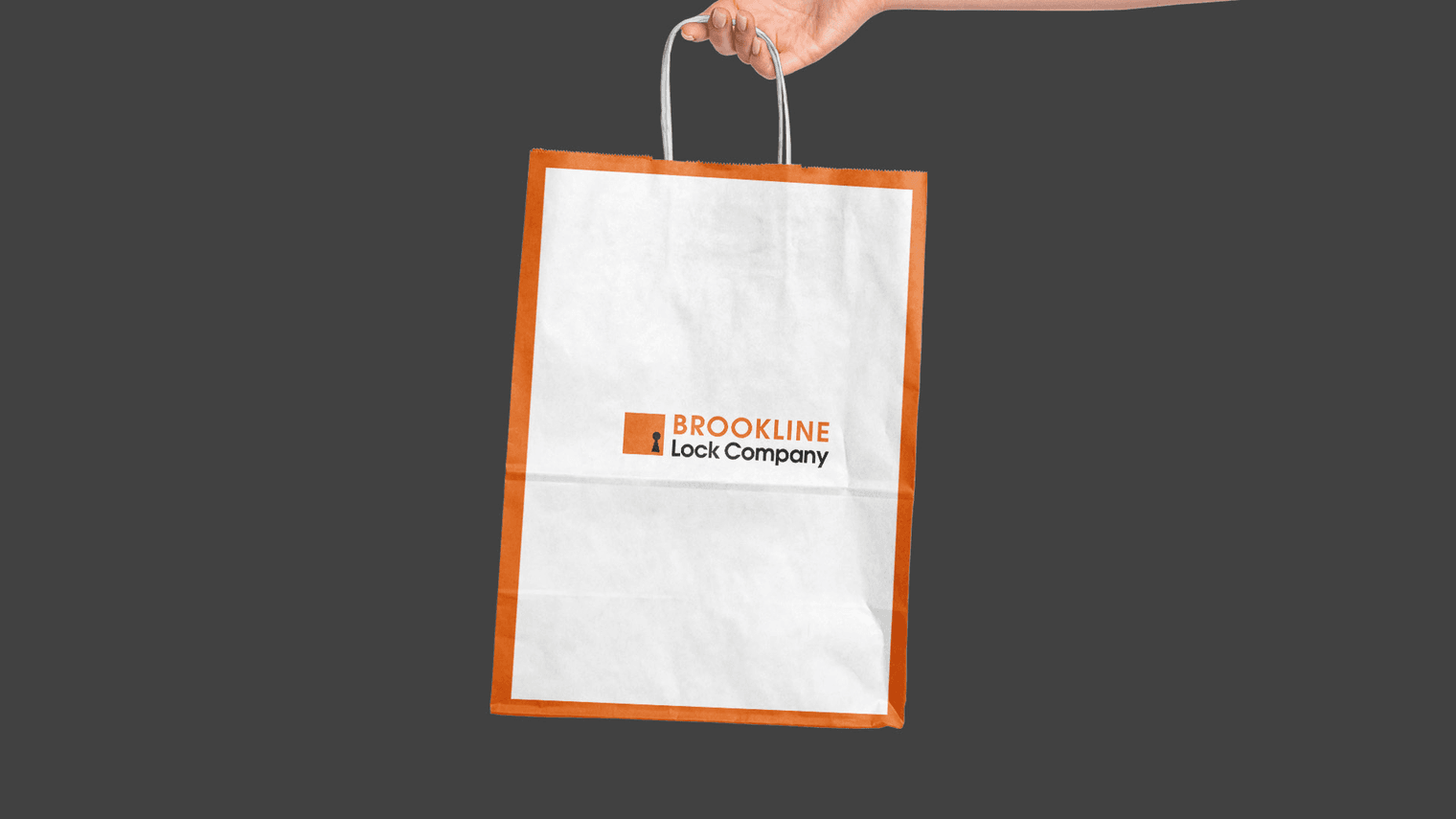 A hand holds a white paper shopping bag featuring an orange frame and the Brookline Lock Company logo against a gray background.