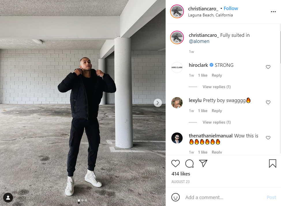 A man in a black jacket and pants stands in a parking garage, posing while adjusting his collar.