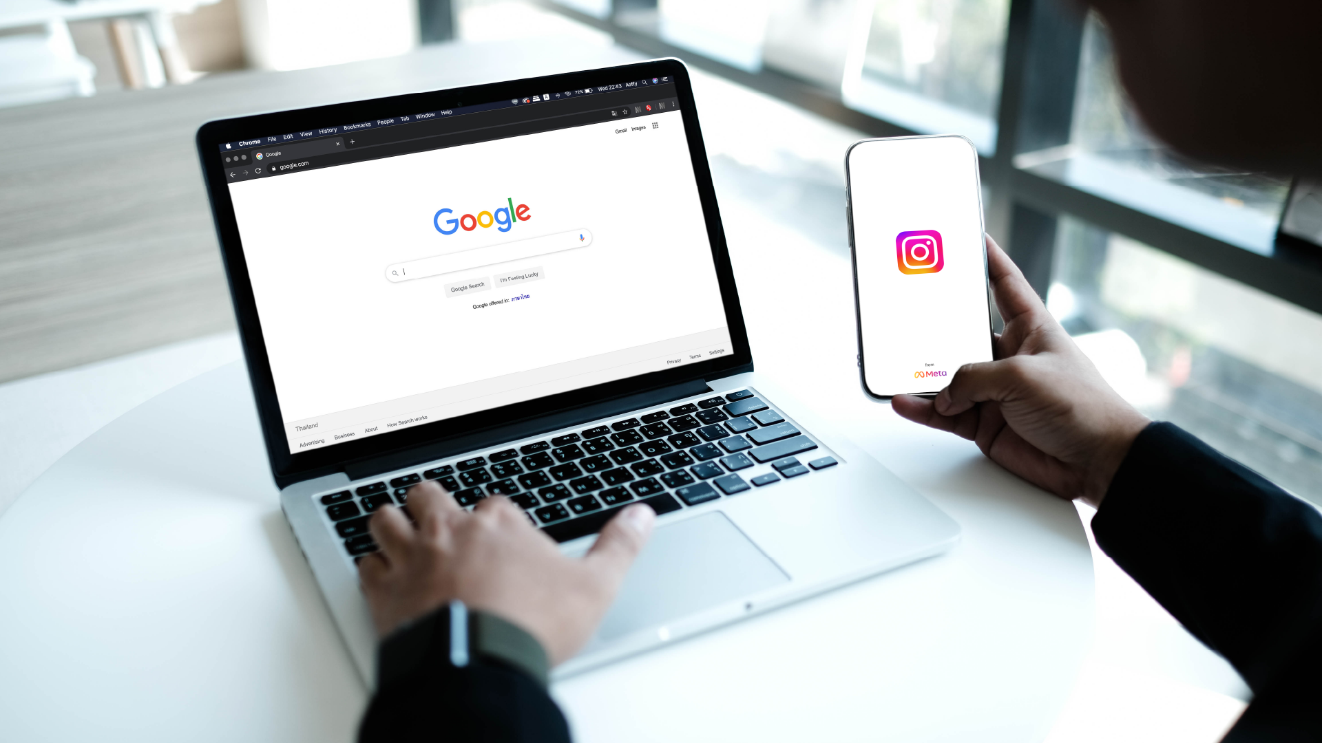 A person sitting at a desk typing on a laptop with Google open, while holding a smartphone displaying the Instagram app.