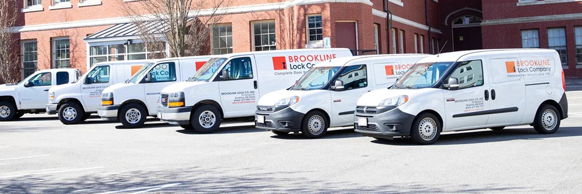 A row of white service vans parked in front of a red brick building on a sunny day.