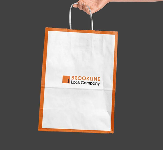 A hand holds a white paper shopping bag featuring an orange frame and the Brookline Lock Company logo against a gray background.