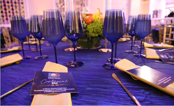 A formal dining table set with deep blue glasses, gold napkins, and event programs on a dark blue tablecloth.
